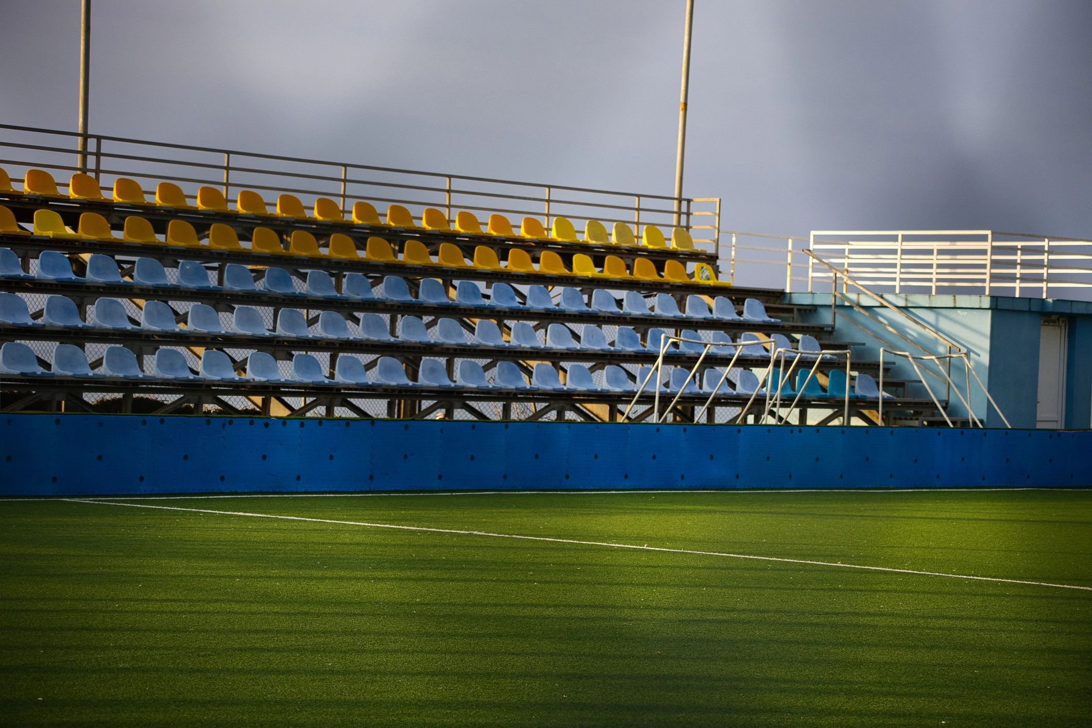 Empty Sports Stadium Bleachers with Yellow and Blue Seats Under Dramatic Sky, Professional Green Artificial Turf Football Field, Outdoor Athletic Arena Infrastructure for Soccer and Events